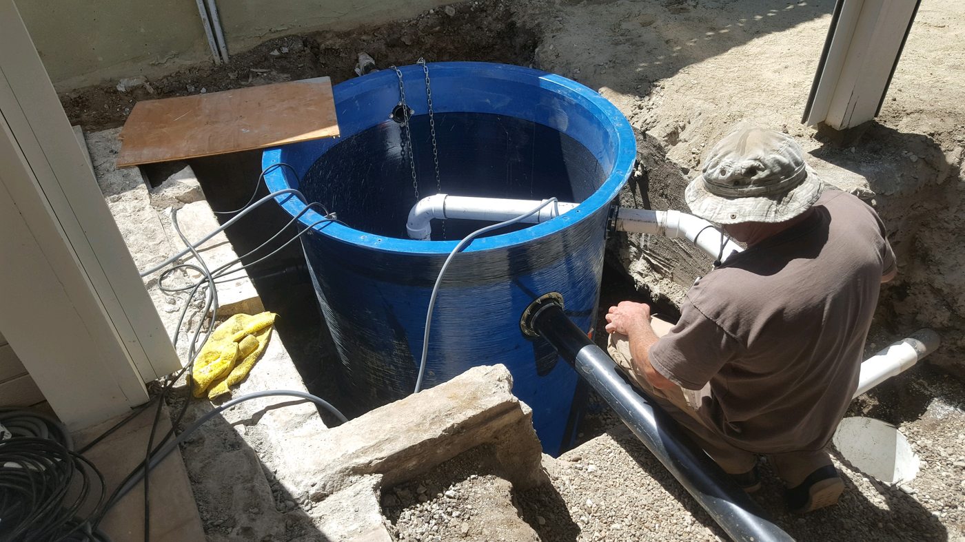 A plumber in a bucket hat kneeling in a trench, connecting pipe to a large blue underground basin.