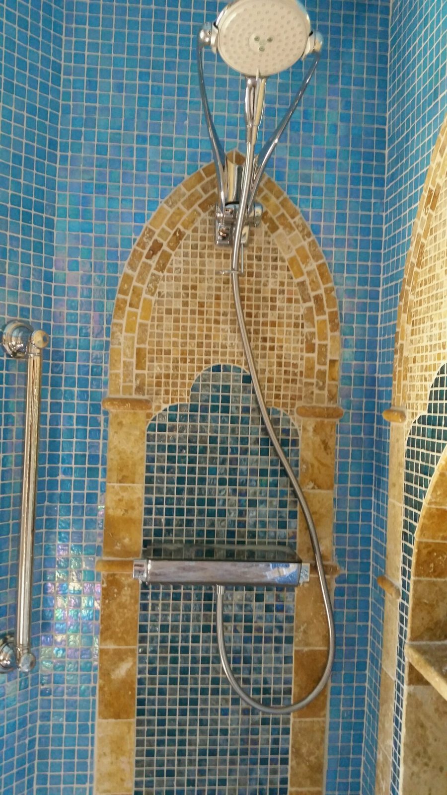A shower with blue glass mosaic walls and gold arches, fitted with a hand shower on a bar and a grab bar.