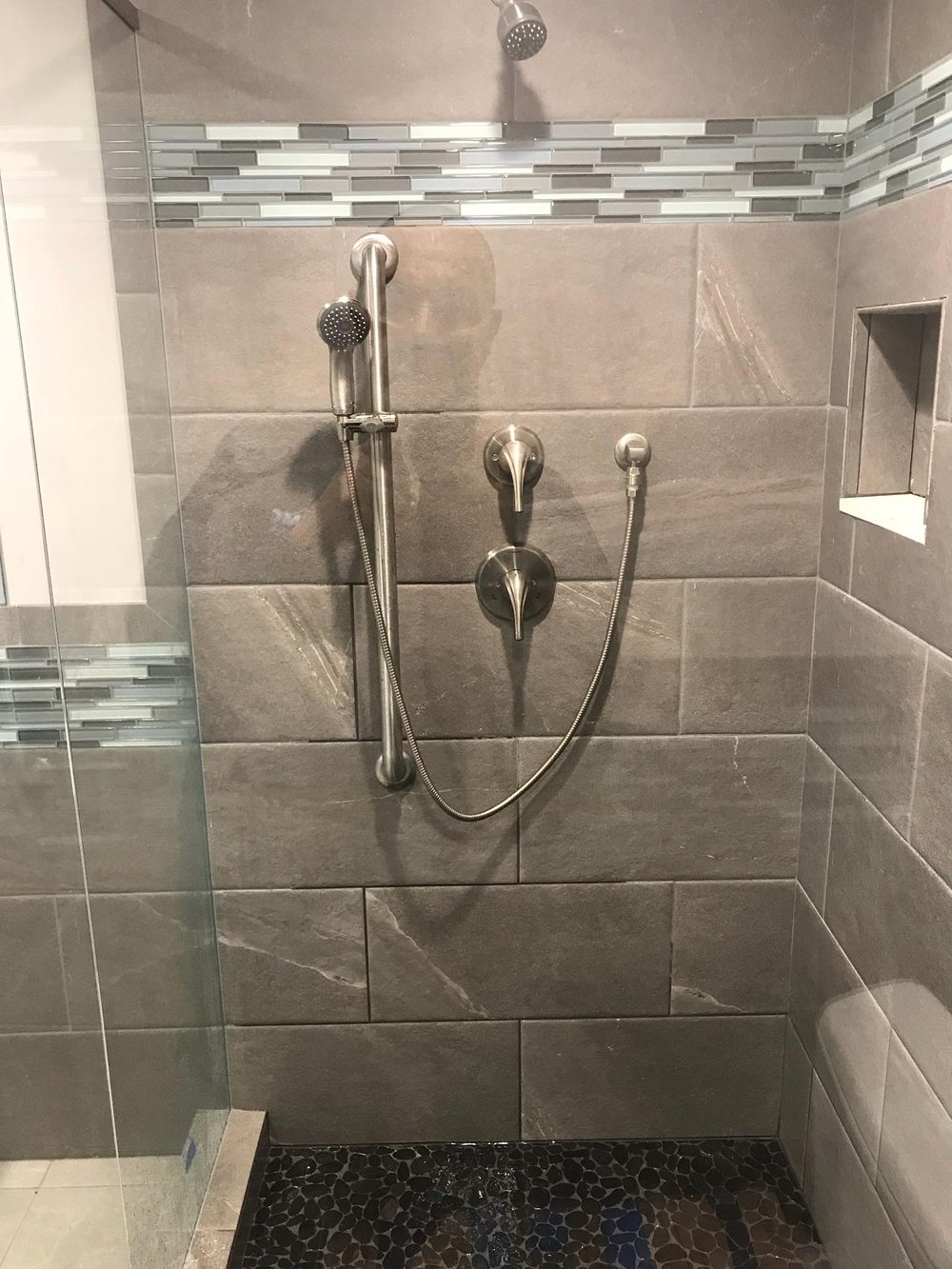 A tiled walk-in shower with a slide-bar hand shower, a valve and a niche, over a pebble floor.