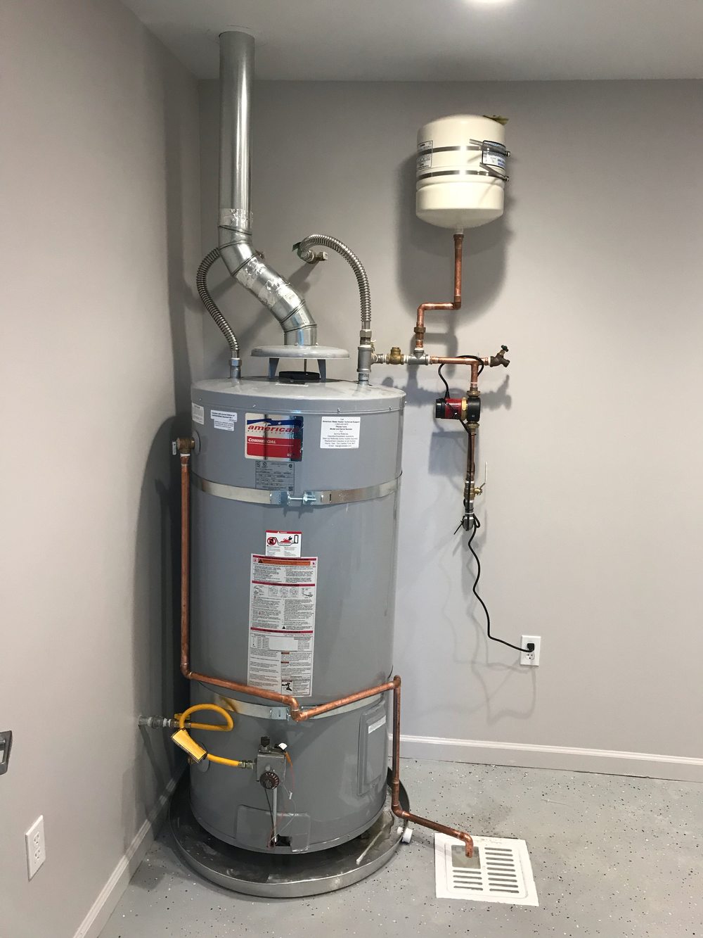 A new gas water heater on a stand in a garage, with copper supply lines, a flexible gas line and an expansion tank above.